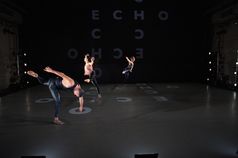 Three dancers in shiny black leggings and tank tops stand on letters that are projected on the floor in off-kilter balances and stances.